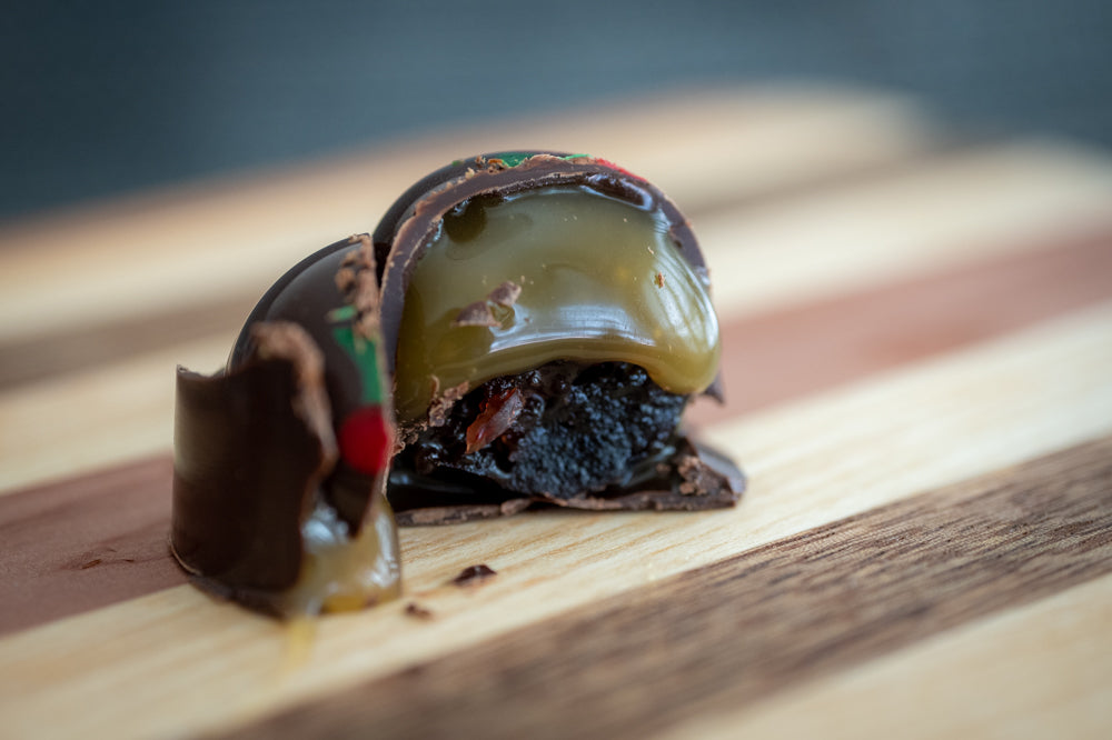 A cut open Candied Cherry & Coconut Milk Caramel Bon Bon oozes caramel sauce onto a wooden cutting board.