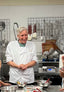 The chocolatier in chef's whites smiles during a kitchen tour and demo, standing at the production kitchen counter beside mixing bowls and ingredients, with tempering tools hanging on the wall behind.