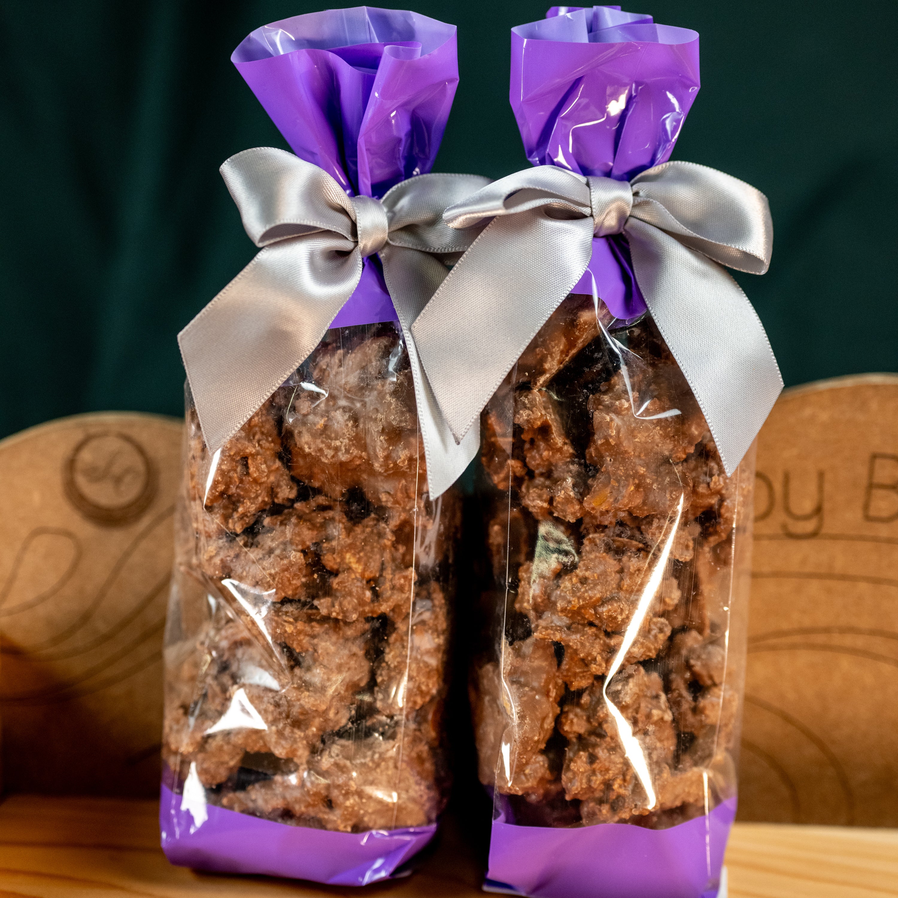 Two clear bags filled with crunchy chocolate pretzel clusters, tied with elegant silver bows and purple tops.