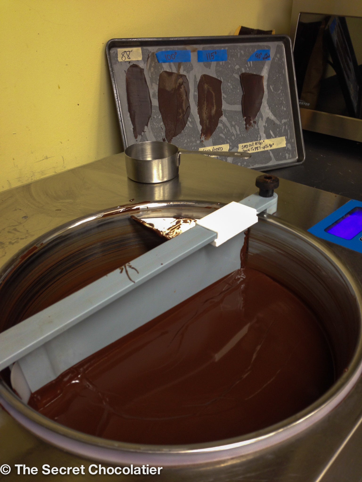 Chocolate tempering machine with a vat of melted chocolate and a test sheet showing different tempering temperatures, used to demonstrate proper technique during The Secret Chocolatier’s bark-making class.