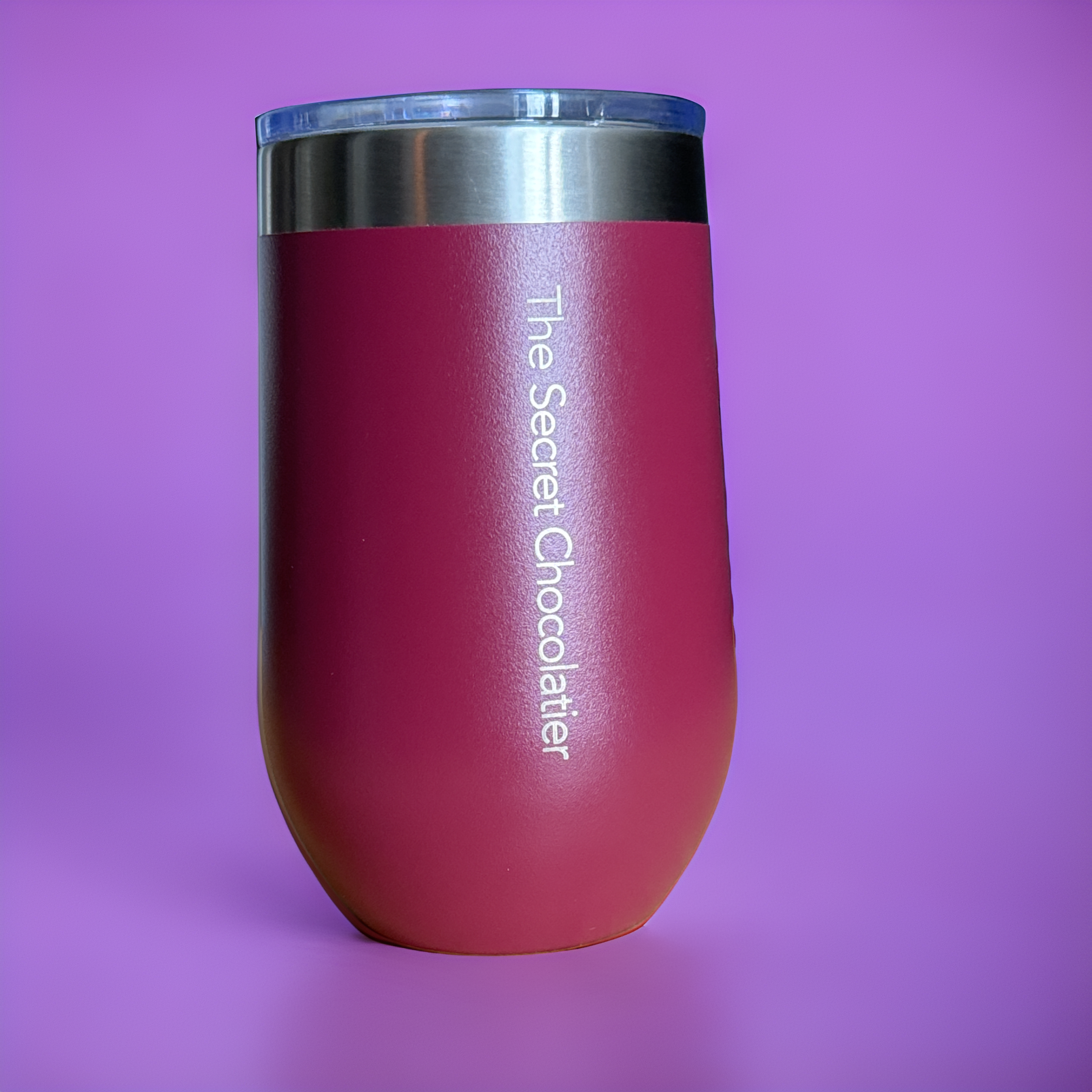 Pink stainless steel insulated tumbler engraved 'The Secret Chocolatier' on the side, product shot against a purple background.