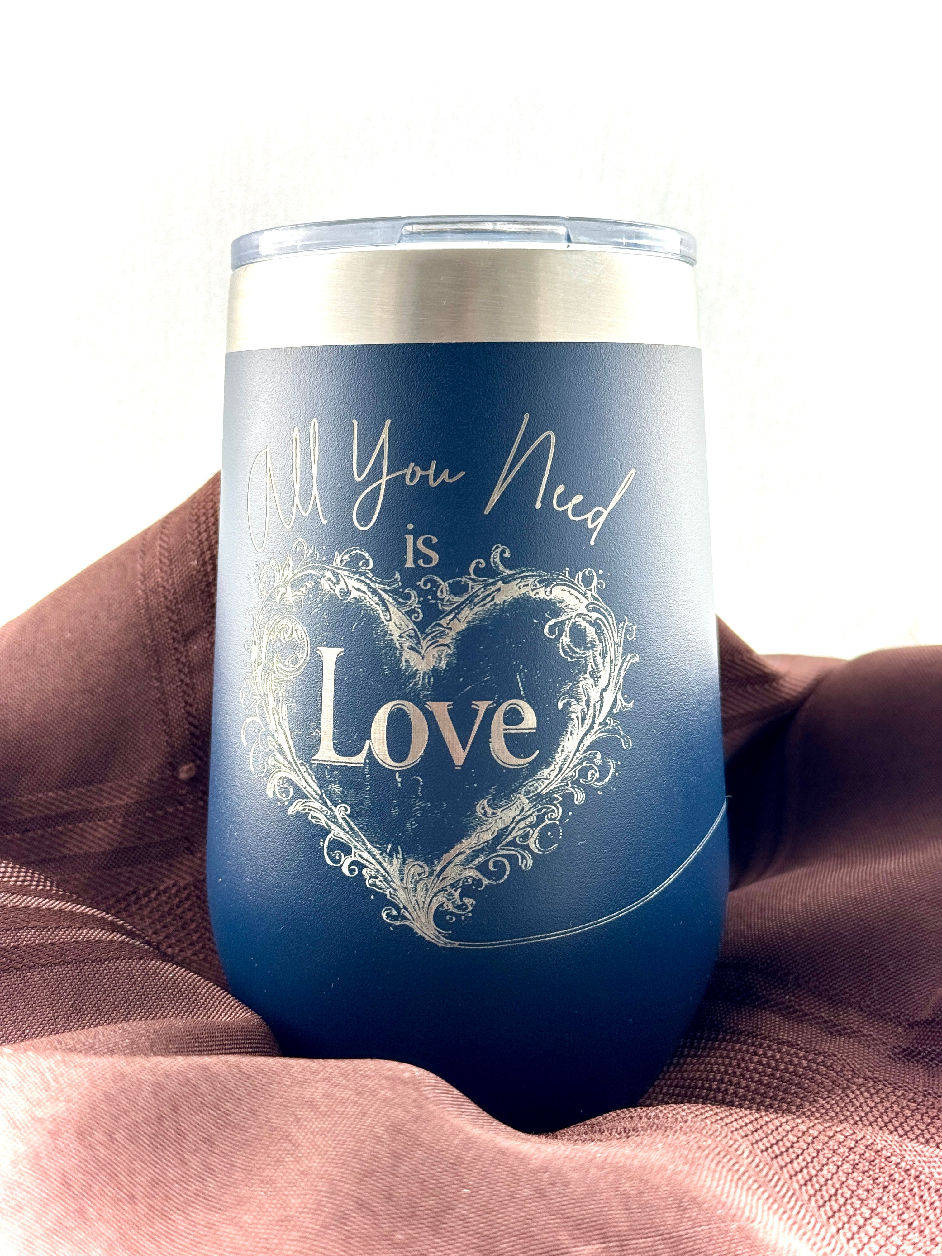 Navy stainless steel tumbler etched with 'All You Need is Love' in script around an ornate heart, displayed on a mauve drape. Main image