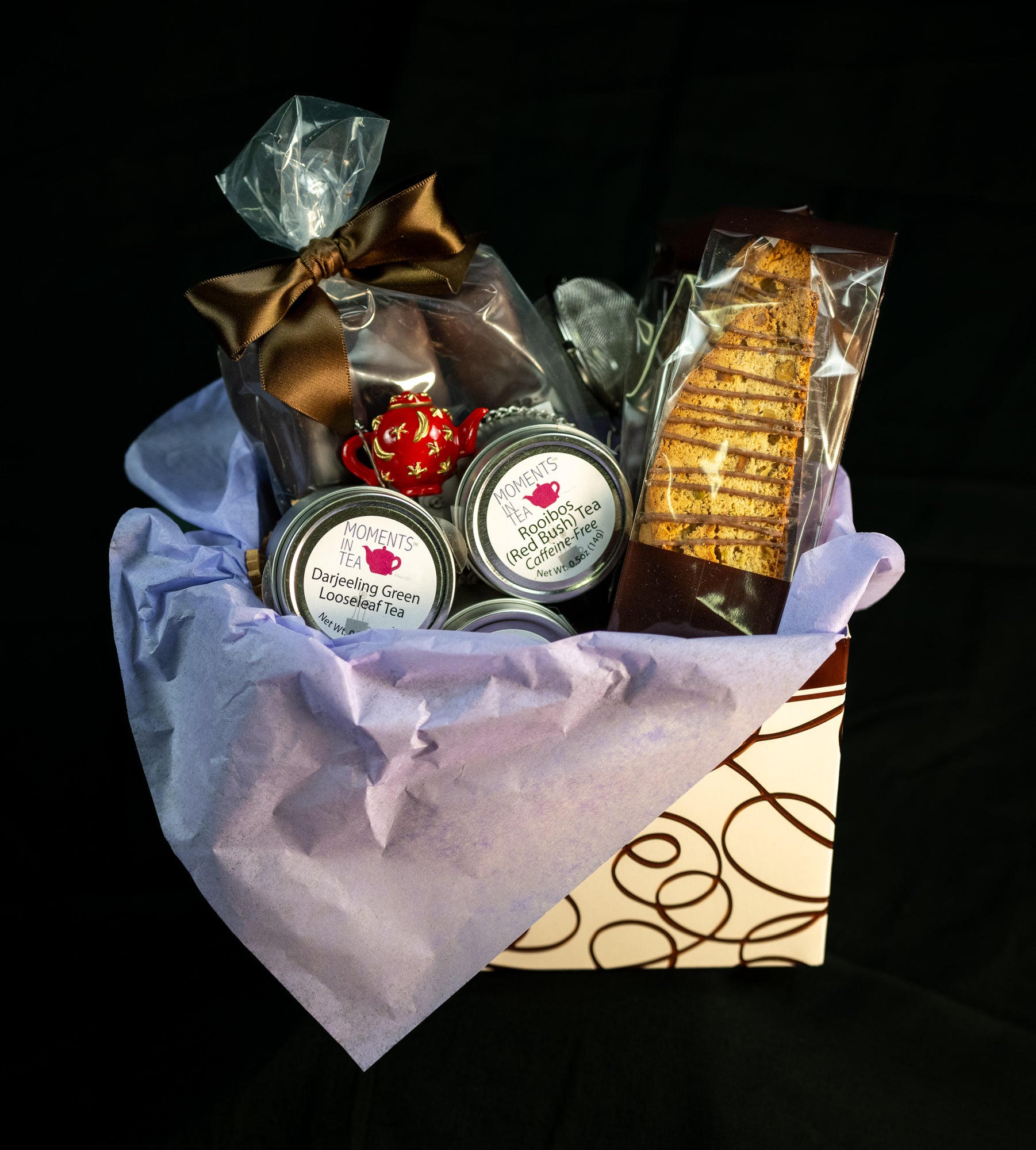 Tea & Chocolate Indulgence Kit — TSC chocolate truffles with brown ribbon, Moments In Tea Darjeeling Green and Rooibos Red Bush tins, biscotti, and a decorative red teapot ornament. Main image