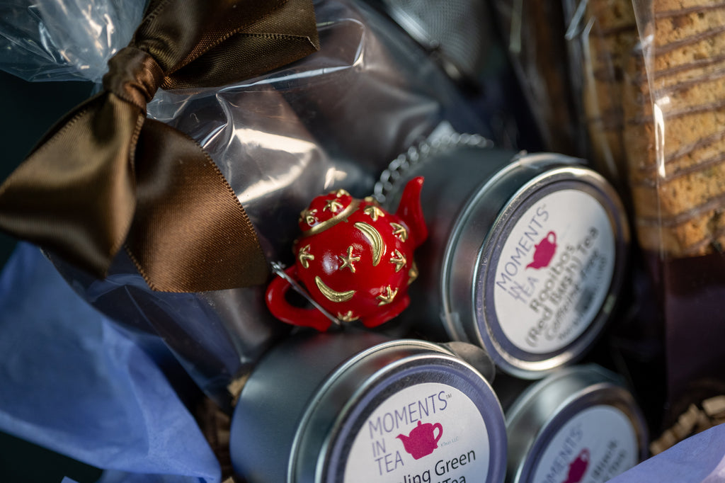 Tea & Chocolate Indulgence Kit detail — Moments In Tea tins and a small red teapot ornament alongside brown-ribbon chocolates.
