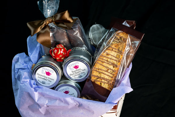 Tea & Chocolate Indulgence Kit — three Moments In Tea tins (Darjeeling Green, Rooibos Red Bush, American Black), biscotti, chocolates with brown ribbon, and a tea strainer.