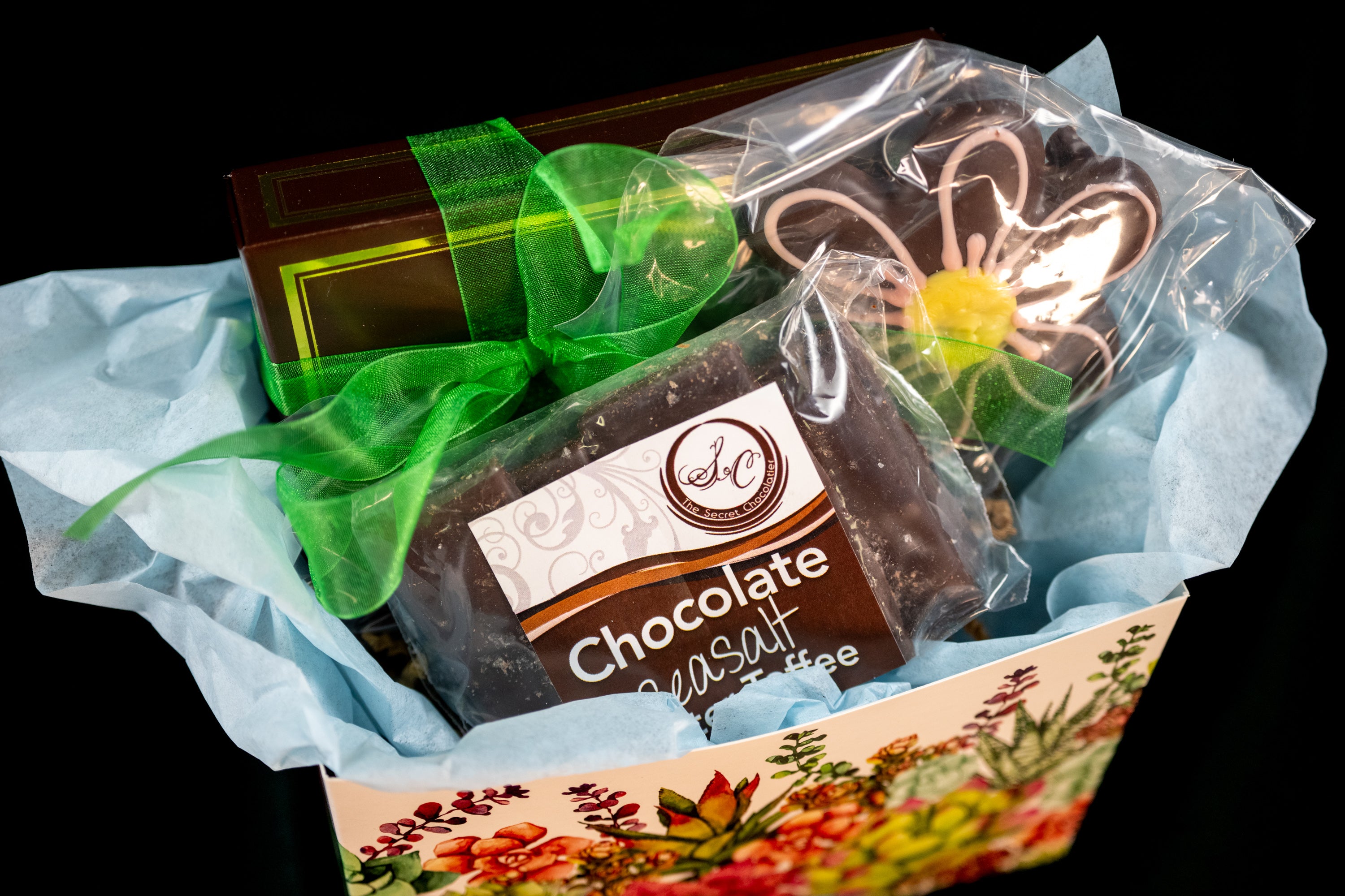 Mother's Day gift box detail — Chocolate Sea Salt Butter Toffee, a chocolate daisy cookie, and TSC chocolates wrapped with a green ribbon in a floral-pattern box.