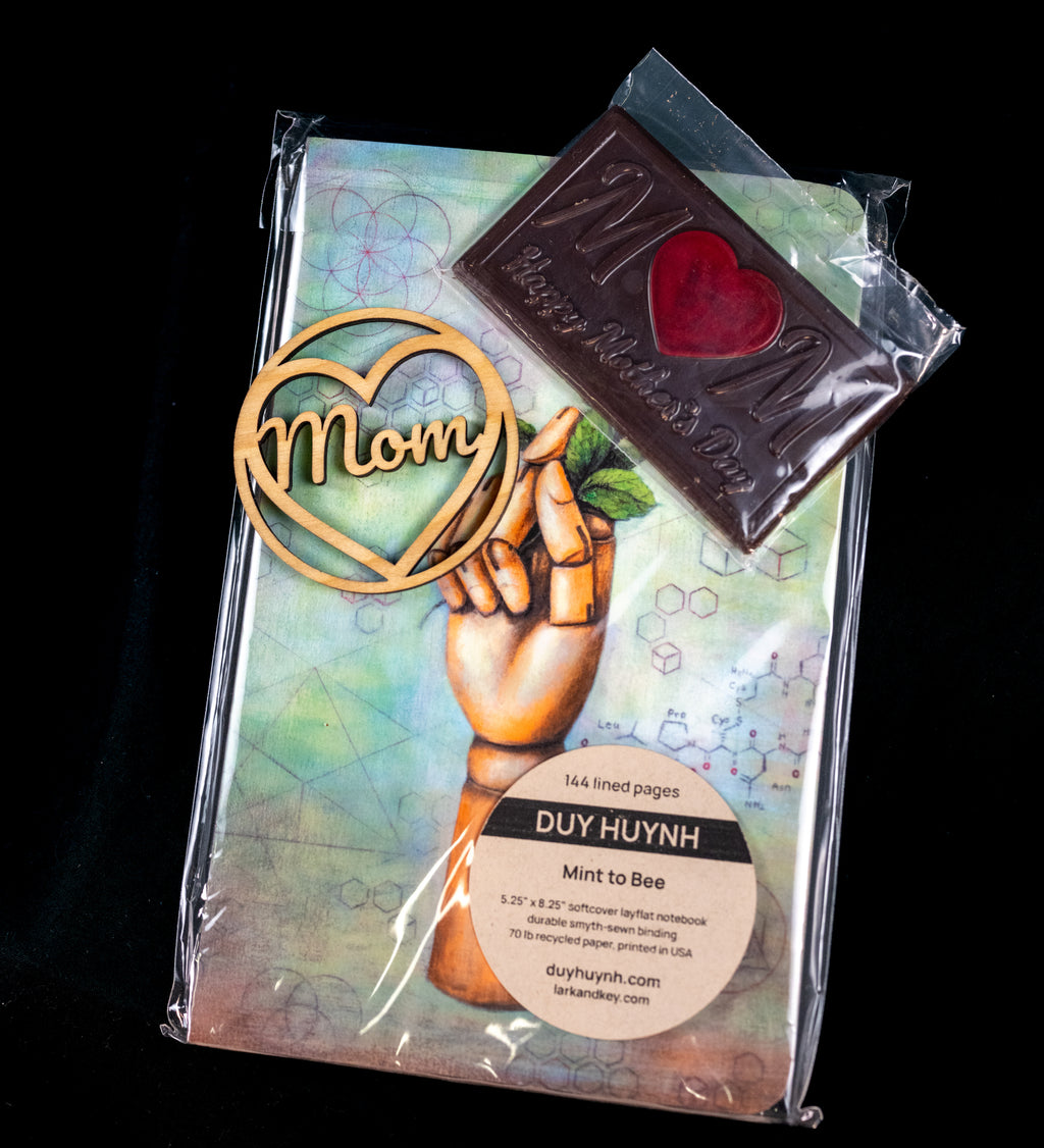Mother's Day gift set — wooden 'Mom' heart ornament, a chocolate M bar with a red heart center, and a Duy Huynh 'Mint to Bee' notebook from Lark and Key.