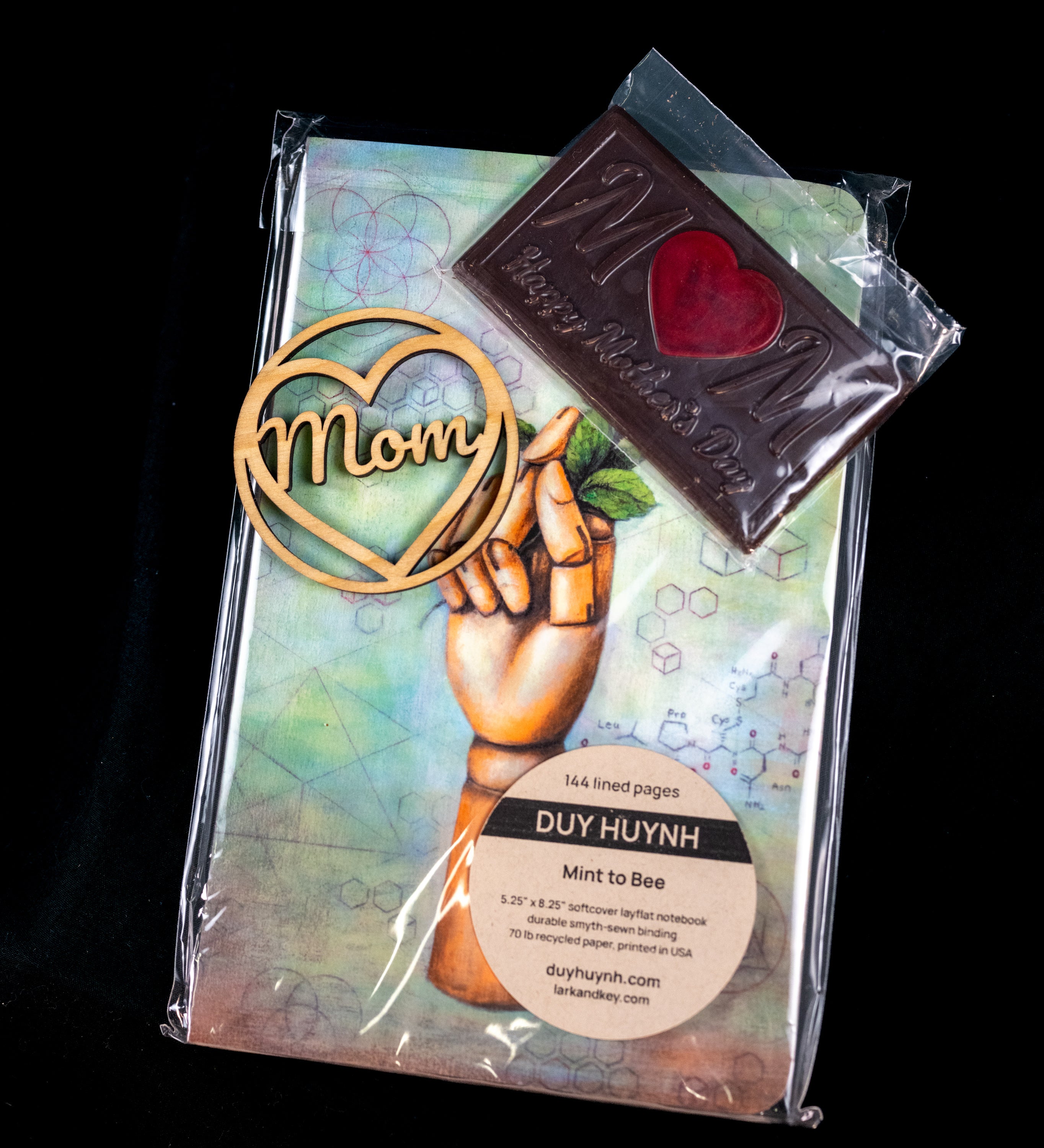 Mother's Day gift set — wooden 'Mom' heart ornament, a chocolate M bar with a red heart center, and a Duy Huynh 'Mint to Bee' notebook from Lark and Key.