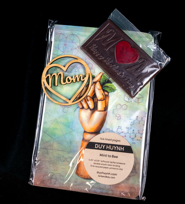 Mother's Day gift set — wooden 'Mom' heart ornament, a chocolate M bar with a red heart center, and a Duy Huynh 'Mint to Bee' notebook from Lark and Key.
