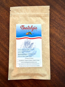 Nostalgia Chocolate Dominican Republic single-origin bar in a kraft-paper bag with a light-blue cacao-pod label — Reserva Zorzal cacao, fig, cherry, and lime zest tasting notes.
