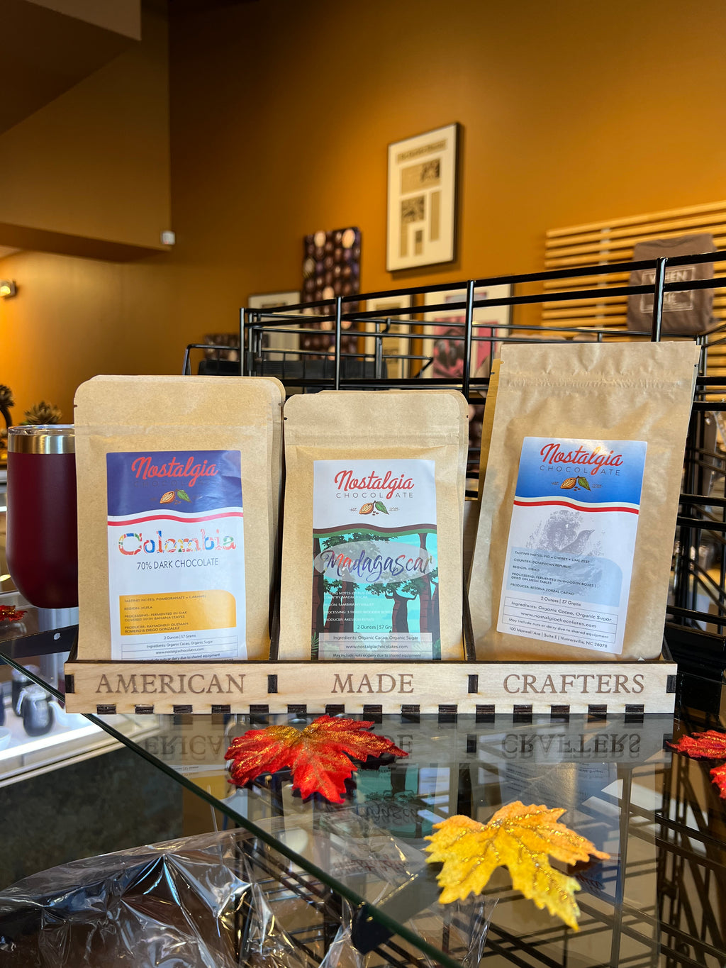 Three Nostalgia Chocolate single-origin bars — Colombia, Madagascar, and Dominican Republic — on a wooden 'American Made Crafters' display rack in the shop, with autumn leaves on the case below.