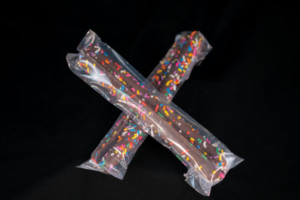 Two cellophane-wrapped Chocolate Pretzel Sticks finished with rainbow sprinkles, crossed on a dark background.