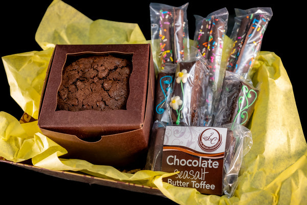 The Secret Chocolatier Snack Box on yellow tissue — a box of Flourless Chocolate Walnut Cookies, a bag of Chocolate Sea Salt Butter Toffee, and chocolate-dipped pretzel sticks with rainbow sprinkles.