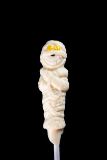 White chocolate mummy lollipop on a stick with hand-piped yellow eyes and a small mouth.