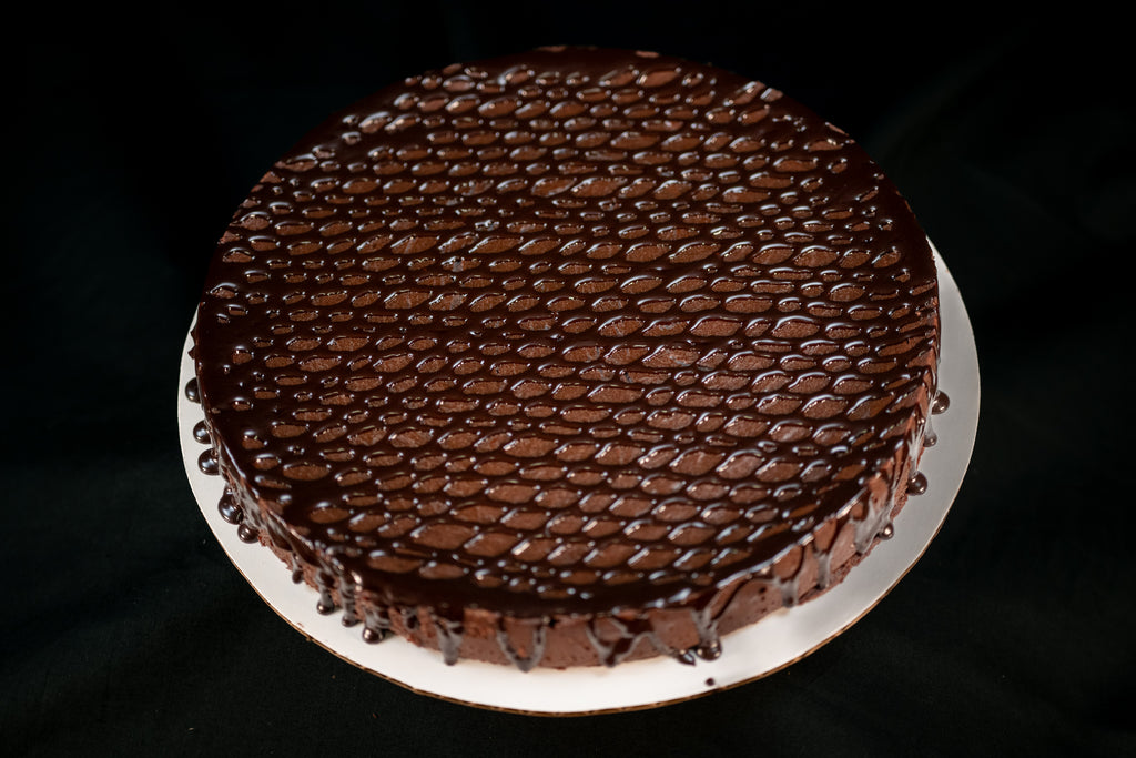 Round Chocolate Oblivion Torte with a glossy quenelle-patterned ganache top and dripping edges, on a white board.