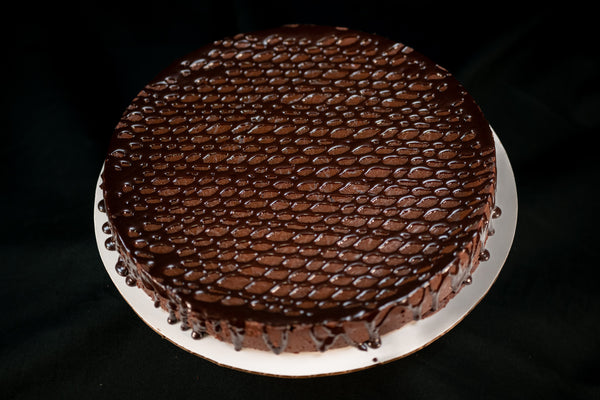 Round Chocolate Oblivion Torte with a glossy quenelle-patterned ganache top and dripping edges, on a white board.