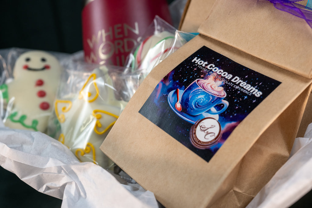 Cozy Cocoa Collection close-up — Hot Cocoa Dreams pouch with a painted hot cocoa label, beside hand-piped white chocolate snowman and angel pieces.