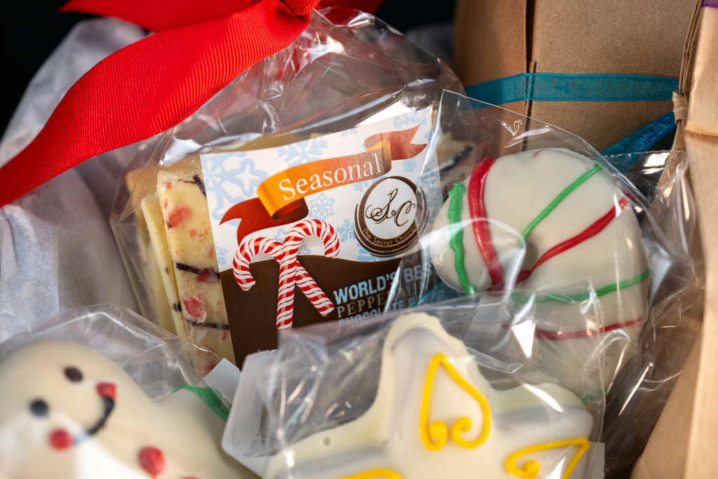 Cozy Cocoa Collection close-up — Seasonal World's Best Peppermint Chocolate Bar with a candy-cane tie, peppermint truffle ball with red and green stripes, and hand-piped snowman and angel chocolates.