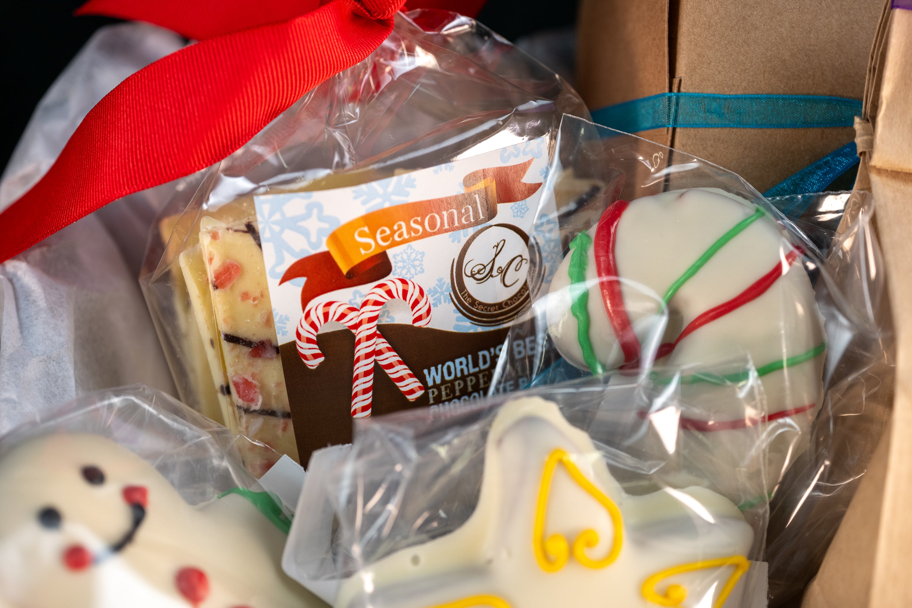 Cozy Cocoa Collection close-up — Seasonal World's Best Peppermint Chocolate Bar with a candy-cane tie, peppermint truffle ball with red and green stripes, and hand-piped snowman and angel chocolates.