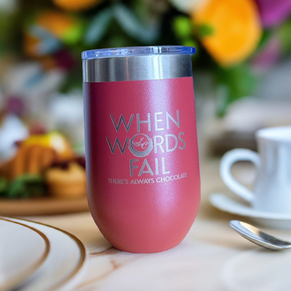 Pink stainless steel insulated tumbler engraved 'When Words Fail There's Always Chocolate', set on a cafe table with pastries.