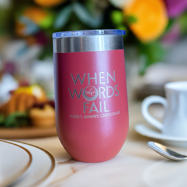 Pink stainless steel insulated tumbler engraved 'When Words Fail There's Always Chocolate', set on a cafe table with pastries.