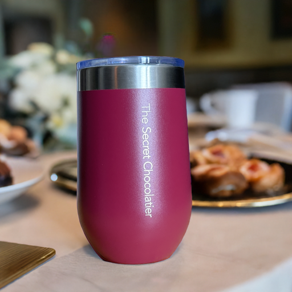 Pink stainless steel tumbler engraved with 'The Secret Chocolatier' vertically on the back, set on a cafe table with pastries.