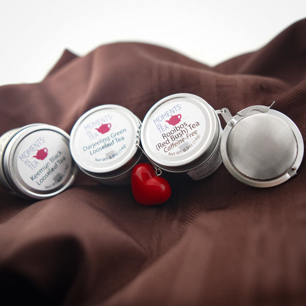 Moments In Tea tins (Keemun Black, Darjeeling Green, Rooibos Red Bush) lined up with a red heart-shaped tea strainer.