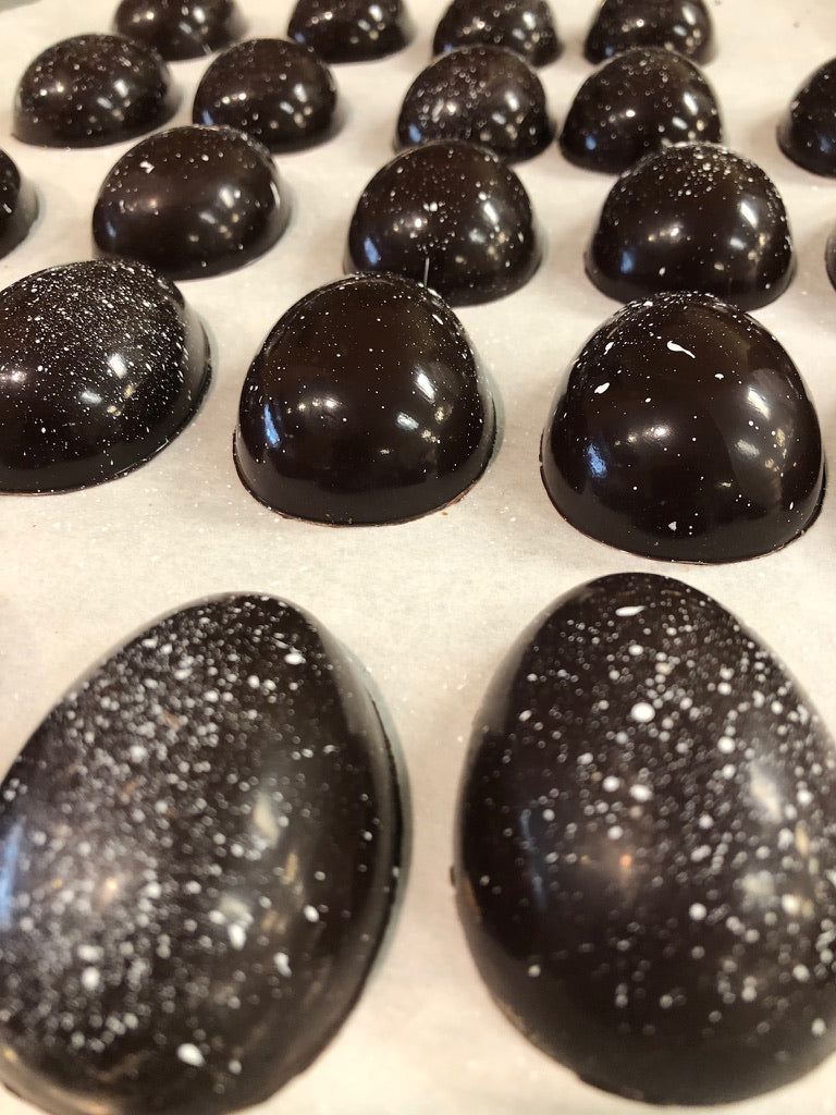 Dark chocolate Easter eggs hand-painted with white luster speckles, in two sizes — small halves and larger full eggs — on parchment paper.