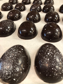 Dark chocolate Easter eggs hand-painted with white luster speckles, in two sizes — small halves and larger full eggs — on parchment paper.