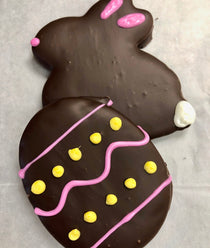 Easter-shaped chocolate-covered graham crackers — a bunny with hand-piped pink ears, nose, and white tail beside an egg with pink ribbons and yellow dot accents.
