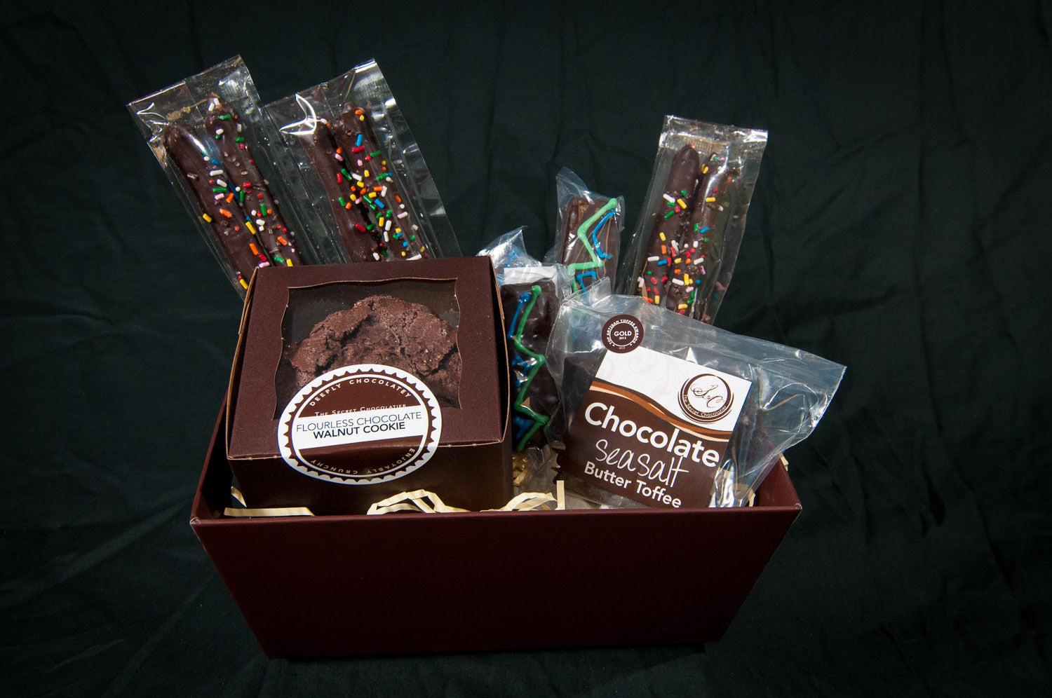 The Secret Chocolatier Snack Box gift basket — Flourless Chocolate Walnut Cookies, Chocolate Sea Salt Butter Toffee, and chocolate-dipped pretzel sticks with rainbow sprinkles and piped accents.