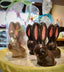 Dark chocolate long-eared sitting bunnies with hand-painted pink ear interiors and pink noses, displayed on a doily — some wrapped in clear cellophane with pastel ribbons.