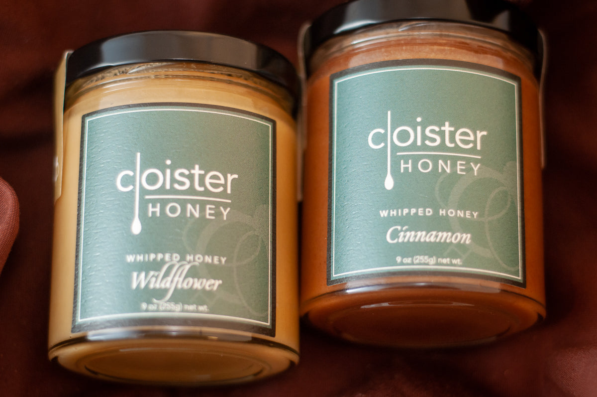 Two Cloister Honey whipped honey jars — Wildflower and Cinnamon — with dark green labels, on burgundy fabric.