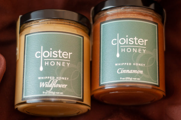 Two Cloister Honey whipped honey jars — Wildflower and Cinnamon — with dark green labels, on burgundy fabric.