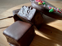 Three chocolate-covered marshmallow squares on a wooden board, with a Valentine's heart piped with floral motifs in the background.