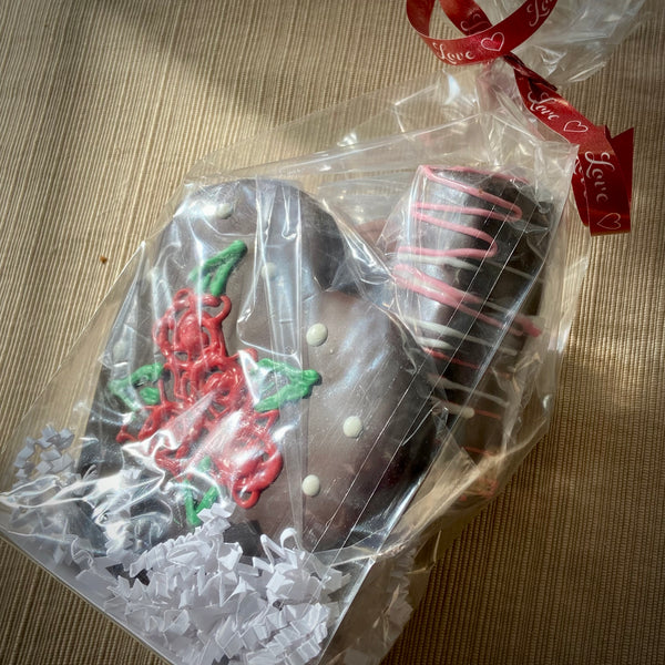 Valentine's Treat Bag — clear cellophane tied with a red 'Love' ribbon — holding a chocolate-dipped heart with piped roses and a chocolate-dipped pretzel stick with pink and white stripes, on white crinkle filler.