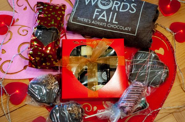 Overhead view of the When Words Fail gift box — TSC shirt with 'WORDS FAIL — THERE'S ALWAYS CHOCOLATE', red heart-window truffle box, chocolate-covered pretzel pop, and heart-shaped chocolate.