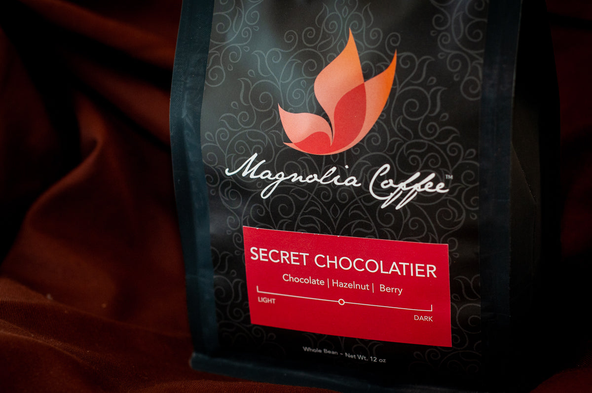 Magnolia Coffee × The Secret Chocolatier whole-bean coffee bag — 12 oz, medium roast with chocolate, hazelnut, and berry tasting notes — on burgundy fabric. Main image