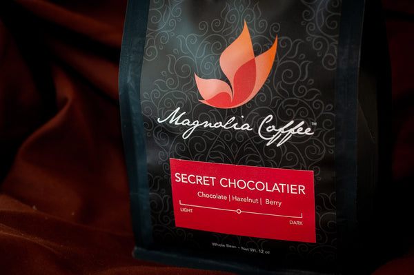 Magnolia Coffee × The Secret Chocolatier whole-bean coffee bag — 12 oz, medium roast with chocolate, hazelnut, and berry tasting notes — on burgundy fabric.