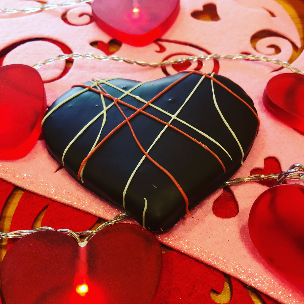 Close-up of a dark chocolate Valentine's heart with red, white, and yellow decorative piping on a pink heart-print background with heart-shaped lights.
