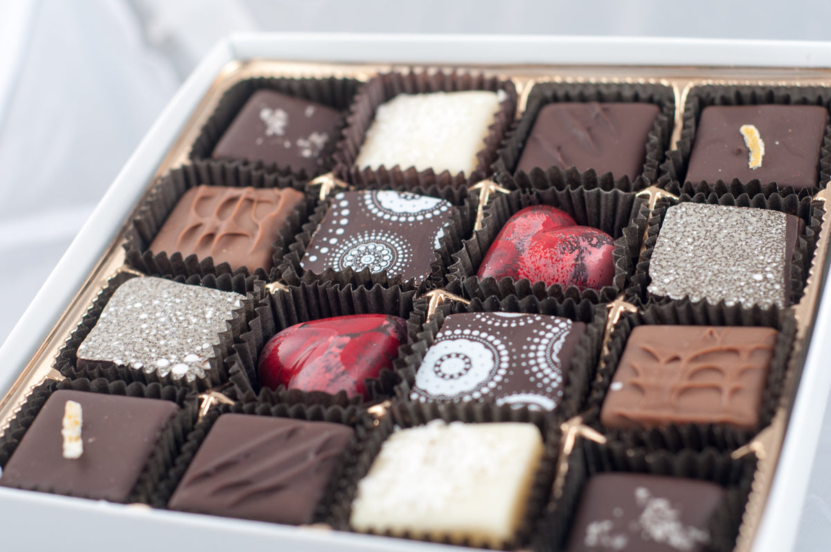 Valentine's Seasonal Variety 16-piece box — red heart-shaped chocolates with painted designs, dark chocolate with silver-sugar sparkle, and white chocolate with citrus accents.