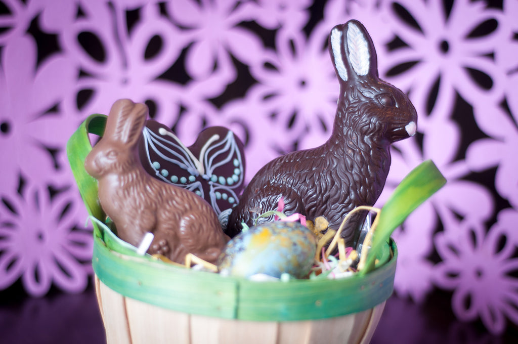 Small Chocolate Easter Basket — wooden basket with a milk chocolate sitting bunny, a piped-icing chocolate butterfly, an upright dark chocolate bunny, and a decorated egg on grass-like filler.