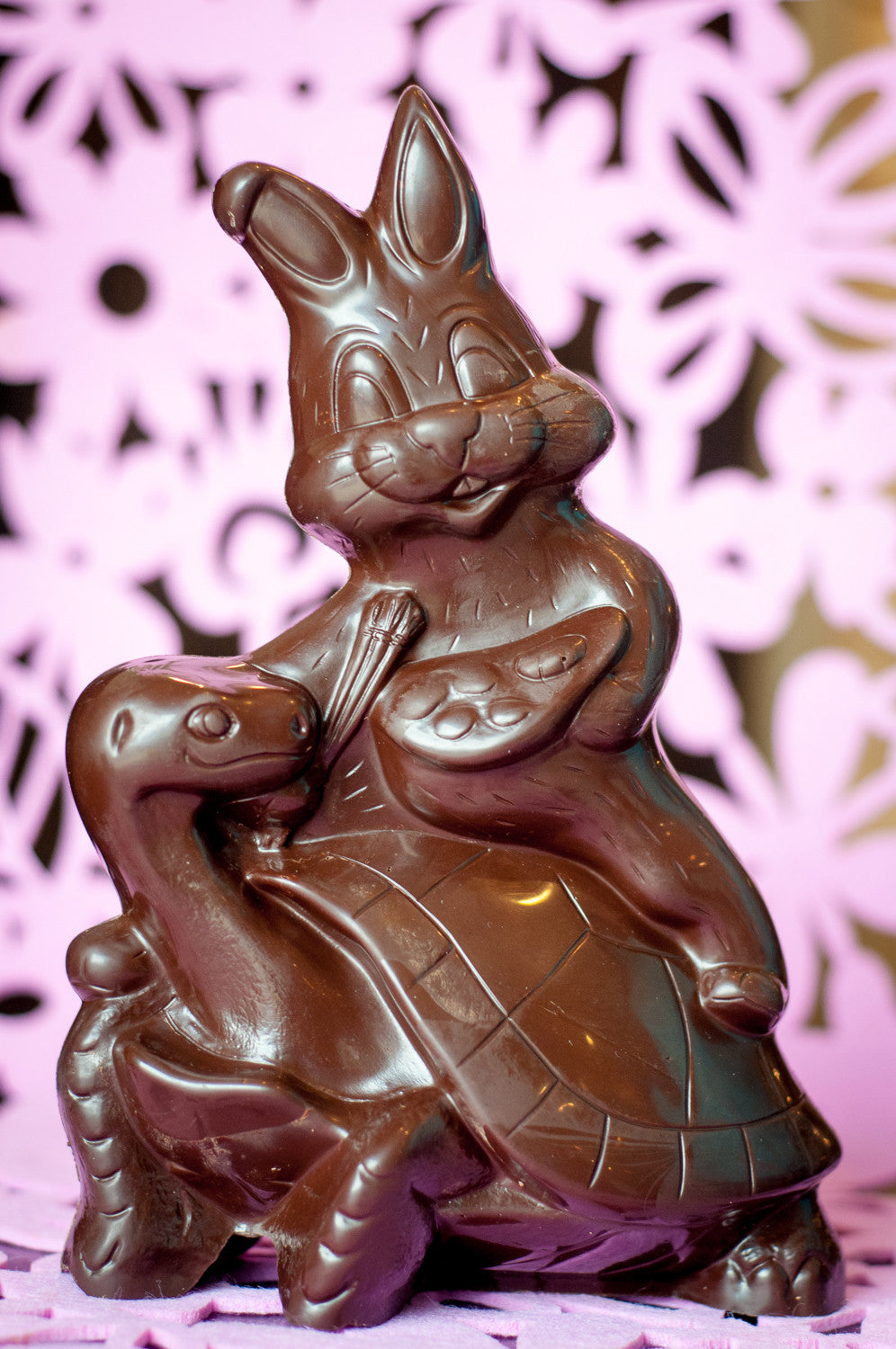 Detailed dark chocolate Easter sculpture of a bunny sitting on a turtle, the bunny holding a carrot. Main image