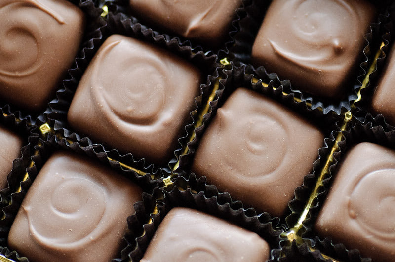 Overhead view of milk chocolate-enrobed caramels in dark paper cups with gold foil peeking through, each finished with a piped chocolate swirl.