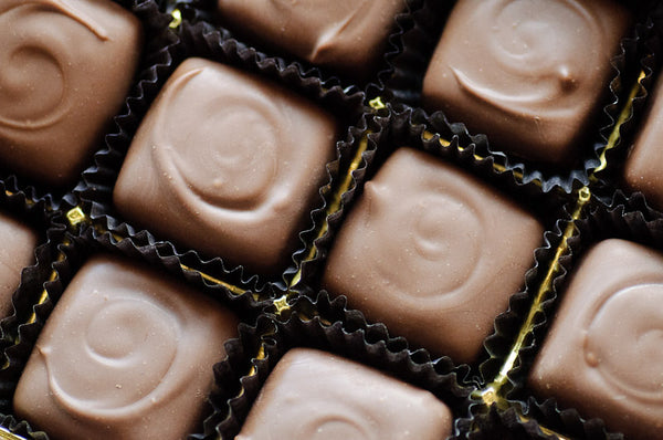 Overhead view of milk chocolate-enrobed caramels in dark paper cups with gold foil peeking through, each finished with a piped chocolate swirl.