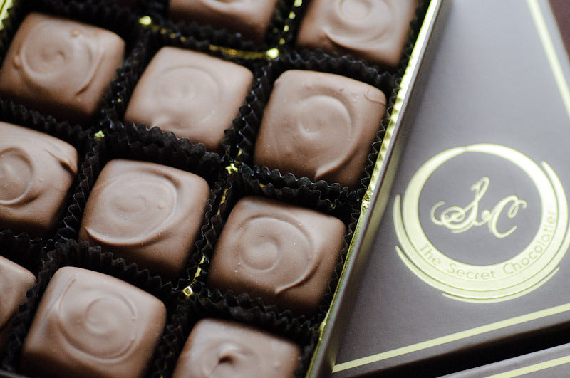Milk chocolate caramels in dark paper cups, alongside The Secret Chocolatier gift box with the gold monogram lid.