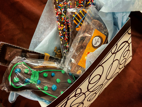 Father's Day gift box opened — chocolate-covered pretzel sticks with rainbow sprinkles, Enrobed Brownies, and a chocolate tie cookie with green and blue dots.