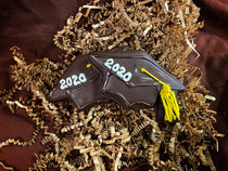 Graduation cap chocolate-covered grahams hand-piped with the class year and a yellow tassel, on brown crinkled paper filler.