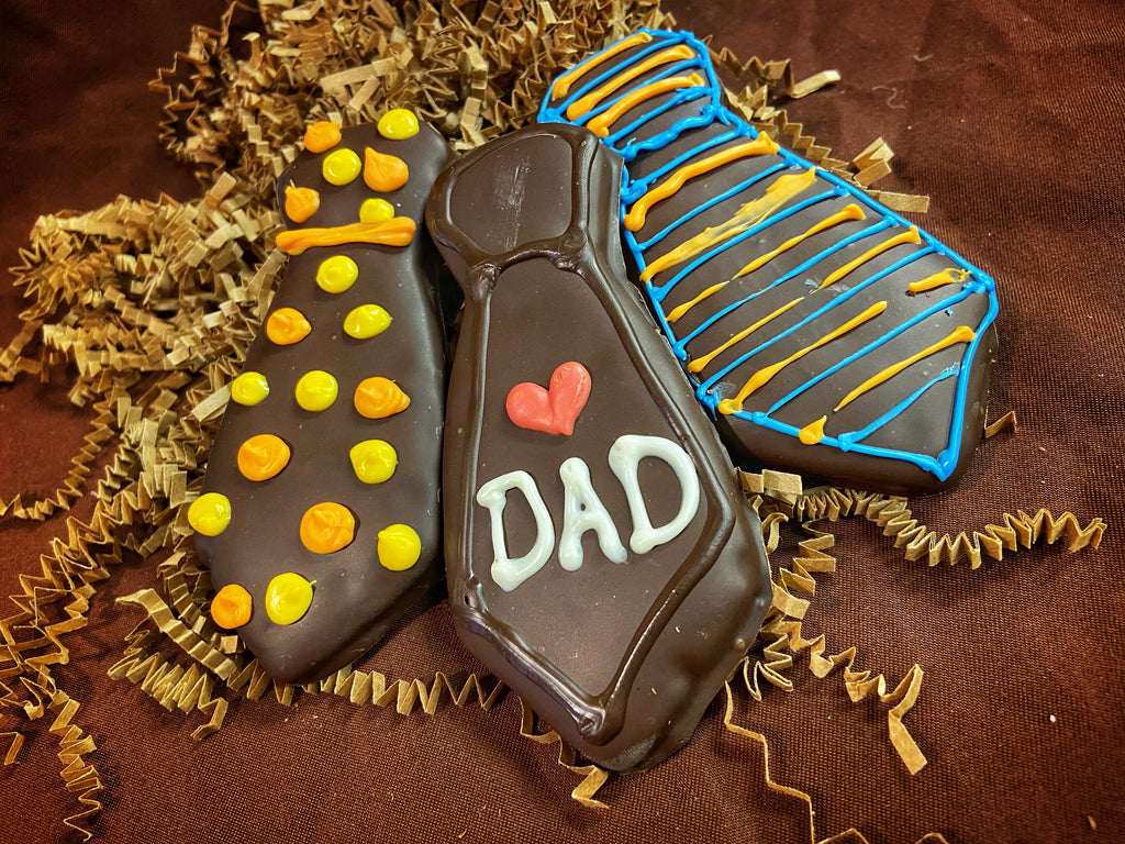Father's Day chocolate-covered graham cracker neckties — one with yellow and orange dot patterns, one piped 'DAD' with a red heart, one with blue and yellow stripes — on crinkled paper filler.