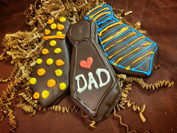 Father's Day chocolate-covered graham cracker neckties — one with yellow and orange dot patterns, one piped 'DAD' with a red heart, one with blue and yellow stripes — on crinkled paper filler.